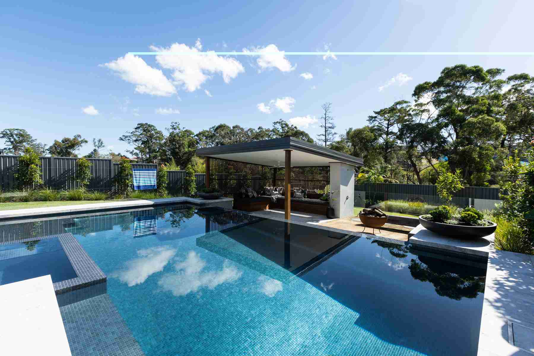 2023 NSW Best Concrete Pool award winner by Splish Splash Pools Pty Ltd, featuring a tropical oasis pool with a matt finish mosaic, generous spa, shallow area, and wet edge in a landscaped garden, complemented by decorative succulent bowls, an outdoor shower, entertaining area, and comfortable day beds shaded by timber screening.